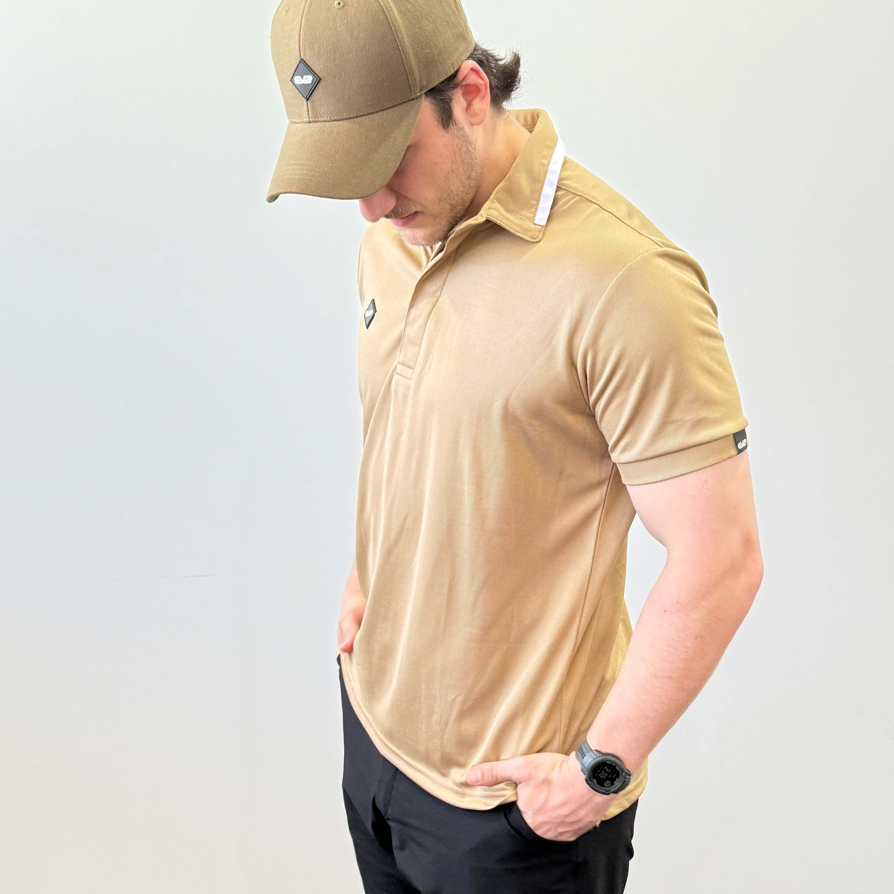 Model wearing EV2 Fairway polo in Camel, left chest has a rubber EV2 logo in the shape of a diamond. Model is wearing EV2 Fairway golf shorts in front of a white wall. 