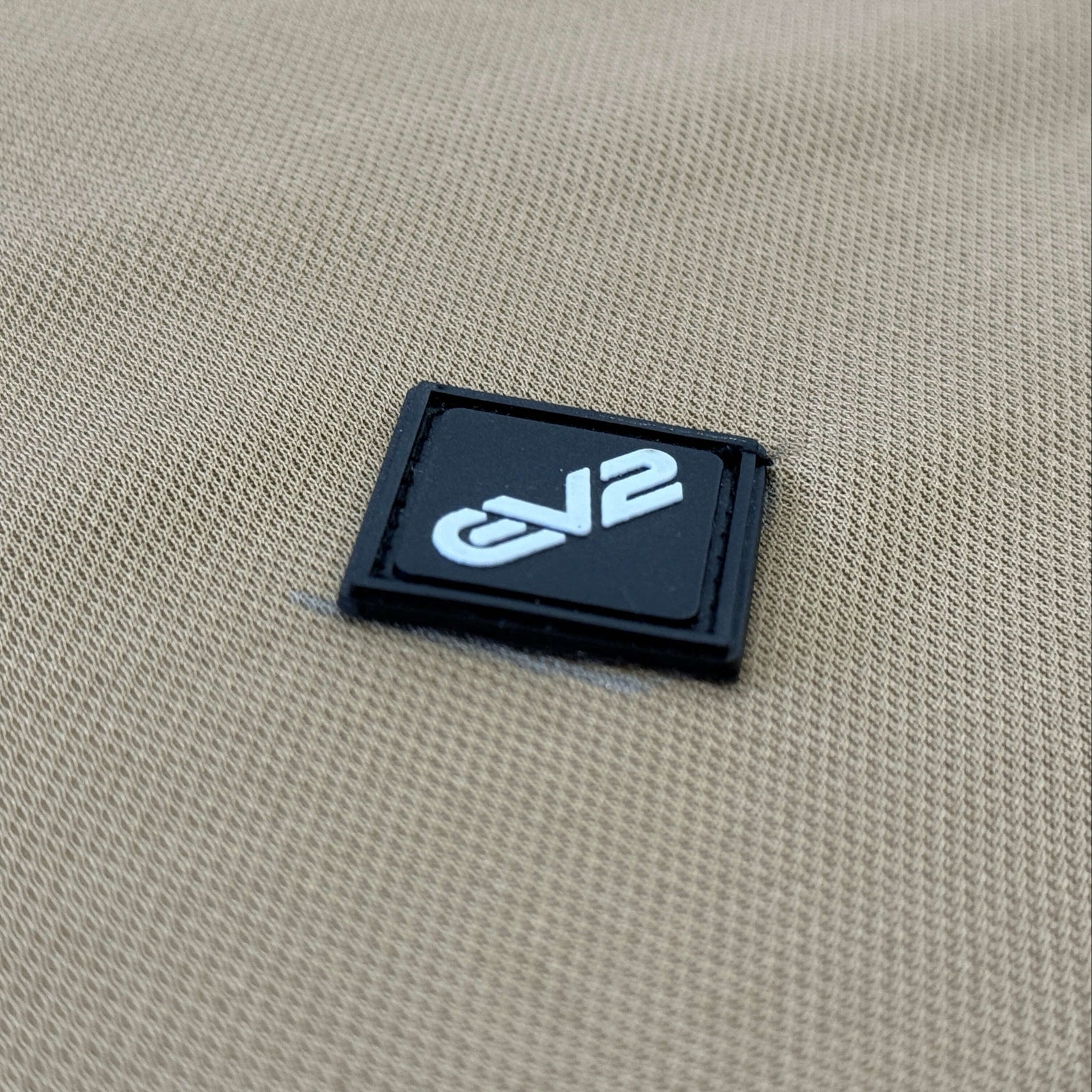 Black diamond shaped rubber patch with 'EV2' logo on a EV2 Fairway Polo in Camel background.